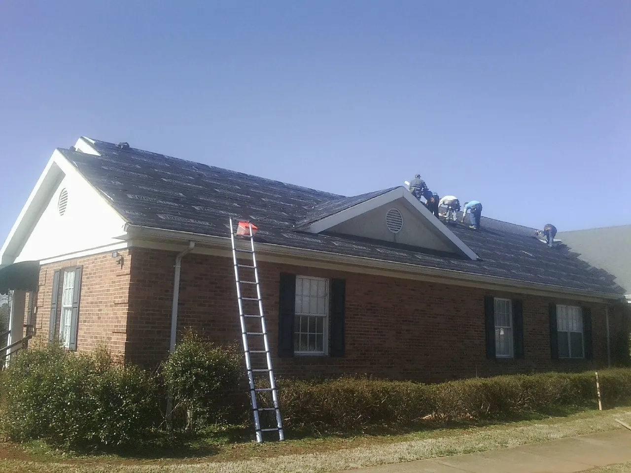 Crew tearing shingles off a brick ranch roof with a ladder set — Metal Roof Repair work by our crew in Croswell