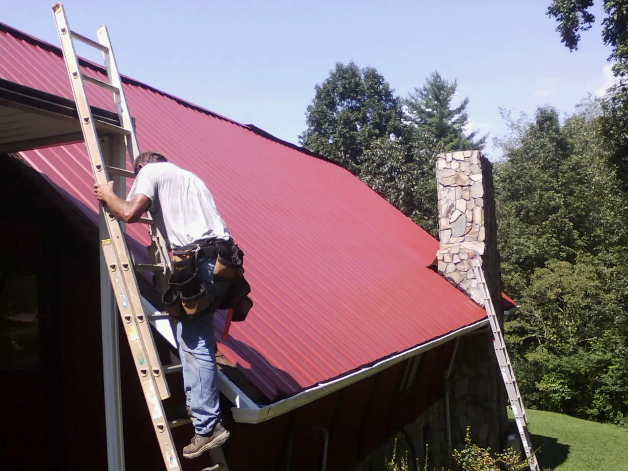 Roofer on a ladder beside a red metal roof and stone chimney — Soffit & Fascia Repair by Roofing Brothers in Croswell